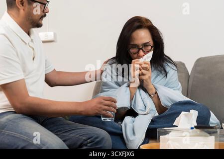 Femme malade enveloppée dans une couverture assise sur un canapé souffrant de rhume et de grippe, tenant le tissu tandis que l'homme offre des soins et un verre d'eau, représentant la maladie Banque D'Images