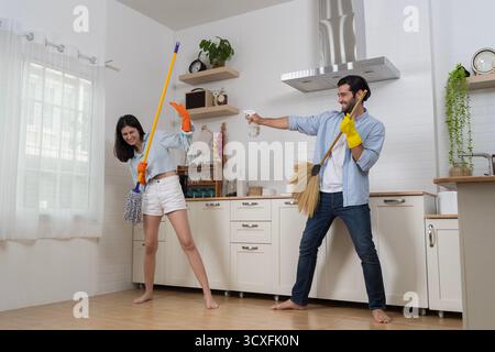 Joyeux couple nettoyant la cuisine ensemble à la maison, en utilisant balai et vadrouille tout en riant et en appréciant les tâches ménagères amusantes, représentant le travail d'équipe, l'amour, Banque D'Images