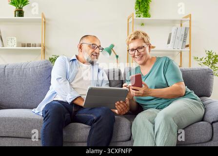 Couple senior utilisant une tablette et un smartphone sur un canapé, apprenant des compétences numériques Banque D'Images