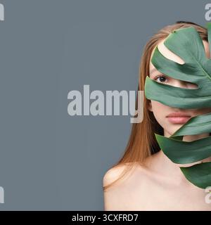Jeune femelle caucasienne partiellement cachée derrière une grande feuille verte sur fond neutre. Banque D'Images