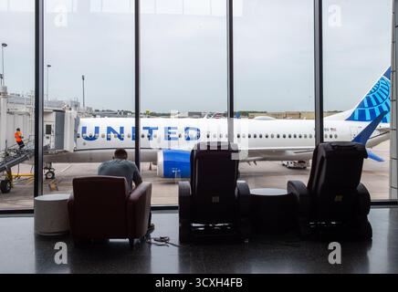 Un voyageur attend près des grandes fenêtres de l'aéroport alors qu'un avion United Airlines se prépare au départ, capturant l'anticipation calme du voyage aérien moderne. Banque D'Images