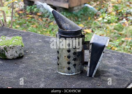 Fumeur d'abeille émettant de la fumée debout sur une surface en béton, équipement d'apiculture avec de l'herbe verte et des feuilles en arrière-plan, à l'extérieur. Banque D'Images