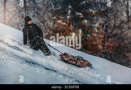 Un garçon vêtu de vêtements chauds d'hiver est assis sur une colline enneigée à côté d'un tube de neige coloré. La lumière du soleil se reflète sur la neige, créant une atmosphère hivernale joyeuse Banque D'Images
