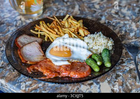 Un copieux petit déjeuner avec œufs frits, jambon, riz, frites croustillantes et cornichons, servi sur une assiette noire décorative. Banque D'Images