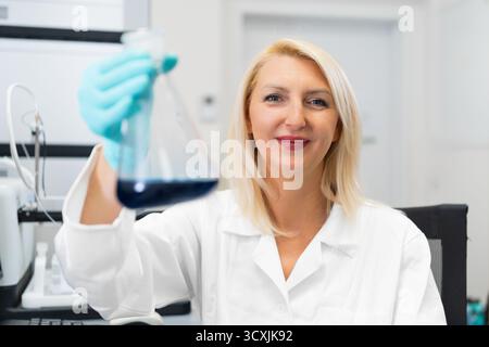 Scientifique femme d'âge moyen en blouse de laboratoire blanche tenant à la solution chimique bleue dans le flacon. Banque D'Images