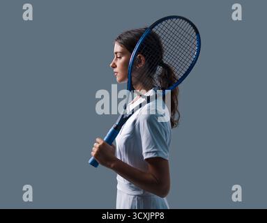Jeune joueur de tennis brune élégant dans une tenue blanche posant avec une raquette sur un fond gris neutre, parfait pour le sport, le fitness, la mode, un Banque D'Images