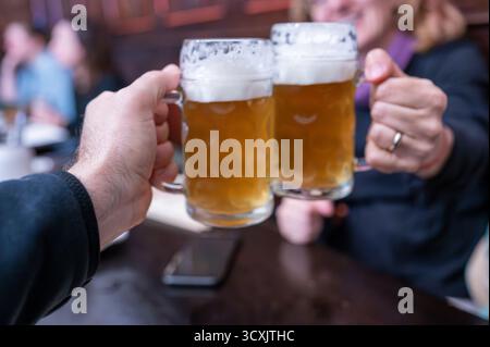 Prague, République tchèque, 7 août 2023. Deux tasses à bière se touchent dans un toast dans un pub. Dans le fond flou, une femme d'âge moyen souriante. Un style de vie o Banque D'Images