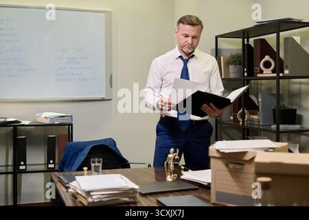 Homme caucasien d'âge moyen debout dans le bureau lisant des documents du dossier, entouré de documents administratifs et de dossiers juridiques, semblant concentré tout en se préparant à une affaire juridique Banque D'Images