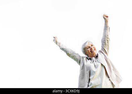 Femme âgée heureuse avec les cheveux gris levant les bras dans la joie, souriant avec confiance sur fond blanc, célébrant la vie, la positivité, la liberté et l'activité Banque D'Images
