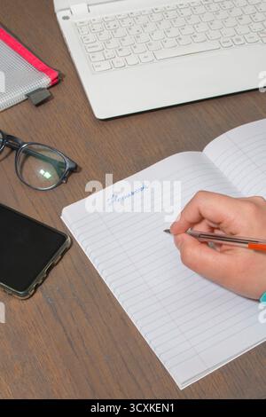 Une personne écrit dans un cahier avec un stylo. Le bloc-notes est ouvert à une page avec le mot devoir écrit dessus. Il y a un téléphone portable sur la table Banque D'Images