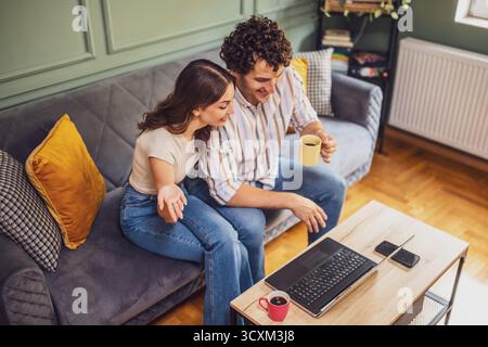 Un couple adulte passe un appel vidéo avec sa famille. Homme et femme assis au canapé à la maison et à l'aide d'un ordinateur portable. Banque D'Images