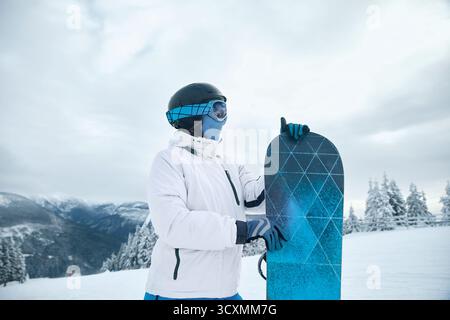 Snowboarder vêtu d'un équipement d'hiver blanc tenant le snowboard sur une montagne enneigée avec des vues panoramiques, prêt pour une aventure de sports d'hiver Banque D'Images