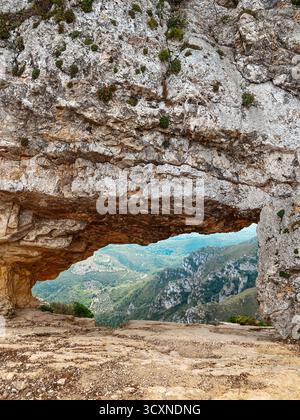 Vue rapprochée de la voûte rocheuse percée connue sous le nom de la Foradada, dans la chaîne de montagnes de la Serra del Montsia, en Espagne, encadrant une vallée lointaine couverte de végétation luxuriante Banque D'Images