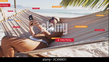 Femme couchée portant maillot de bain, lunettes de soleil et bandeau, tenant smartphone dans un hamac à la plage Banque D'Images
