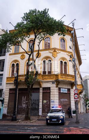 Un policier se tient à côté d'une voiture de patrouille au coin d'une rue, surveillant près d'un bâtiment abandonné et arboré et d'un arbre solitaire par une journée nuageuse Banque D'Images