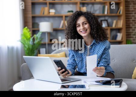 Femme souriante tenant une facture papier, utilisant un smartphone et un ordinateur portable pour gérer son budget personnel et sa planification financière dans un cadre confortable à la maison Banque D'Images