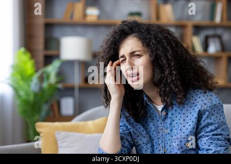 Jeune femme avec les cheveux bouclés grimacant et tenant sa tempe à la maison, souffrant d'un mal de tête douloureux ou de migraine et se sentant malaise, stressée et e. Banque D'Images