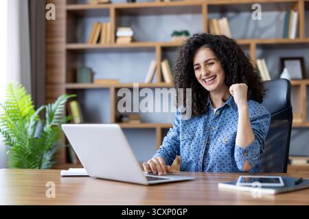 Heureuse femme aux cheveux bouclés célèbre le succès du travail à distance sur un bureau à domicile lumineux, souriant à son ordinateur portable avec joie et une énergie confiante et productive Banque D'Images