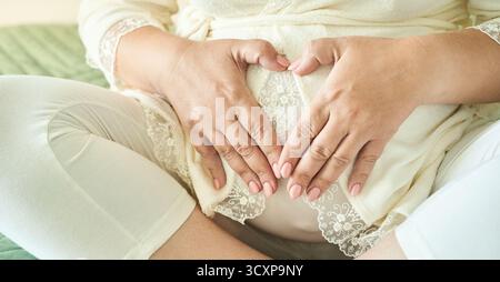 Femme caucasienne enceinte dans la lumière douce formant la forme de coeur sur le ventre dans un cadre confortable. Banque D'Images