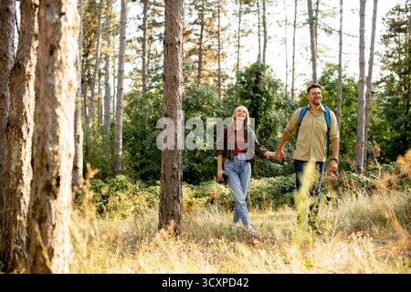Un couple joyeux marche main dans la main sur un sentier forestier serein, entouré de grands arbres et de lumière chaude du soleil. Banque D'Images