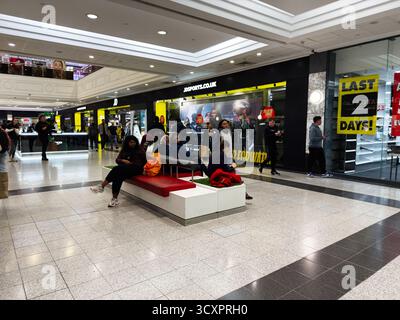Manchester, Royaume-Uni - 26 septembre 2025 : les gens sont assis sur un banc rouge tout en marchant et en faisant du shopping dans un atrium animé du centre commercial Arndale. Banque D'Images