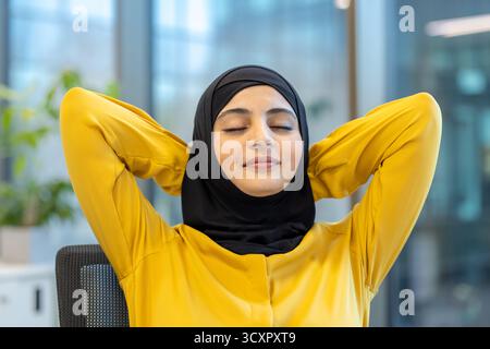 Femme d'affaires musulmane prend une pause paisible à son bureau d'entreprise en se penchant en arrière dans sa chaise les yeux fermés, profitant d'un moment de calme et de s. Banque D'Images