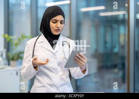 Médecin musulman portant un hijab et un stéthoscope regardant une tablette numérique avec une expression inquiète ou confuse, debout dans un cabinet médical e. Banque D'Images