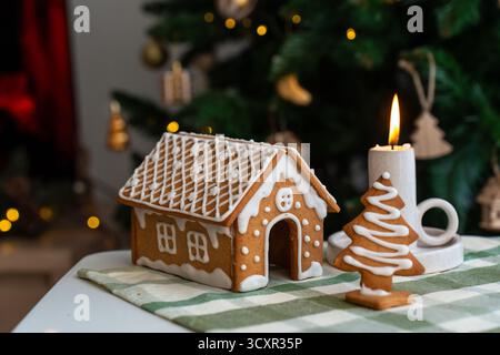 Maison en pain d'épice décorée avec glaçage détaillé et biscuit d'arbre de Noël à côté de la bougie allumée sur la table de fête avec des lumières bokeh. Pâtisserie des fêtes arc Banque D'Images
