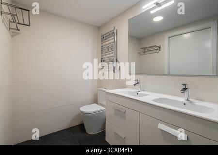 Salle de bains moderne avec lavabo double, grand miroir et toilettes. Palette de couleurs neutres avec des luminaires élégants et un design minimaliste. Banque D'Images