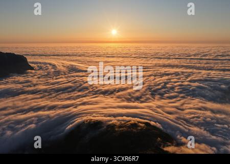 Le soleil se lève au-dessus d'une épaisse couche de nuages ressemblant à un océan, créant une vue aérienne époustouflante des montagnes jetant un œil à travers la brume dorée à l'aube Banque D'Images