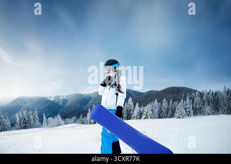 Snowboarder se tient sur une pente enneigée, portant un casque et des lunettes, avec des montagnes majestueuses et des arbres givrés en arrière-plan sous un ciel bleu. Banque D'Images