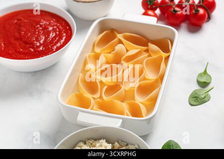 Plat de cuisson avec des pâtes de conchiglie crues et des ingrédients sur fond clair Banque D'Images