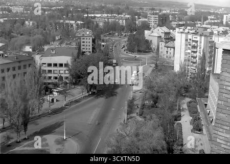 Une photo d'archives panoramique à grand angle des années 1980 capture une scène urbaine animée à Sloviansk, en RSS d'Ukraine. La vue donne sur le carrefour où la rue Dobrovolskoho fusionne avec la rue Yunykh Kommunarov (maintenant rue Bankivska). La large avenue bordée d'arbres est remplie de voitures classiques de l'ère soviétique et d'un fourgon UAZ 'Bukhanka', mettant en valeur la circulation typique de la ville. Cet instantané du mouvement quotidien est un document historique précieux, préservant le rythme ordinaire de la vie dans un Donbass pacifique avant la guerre Banque D'Images