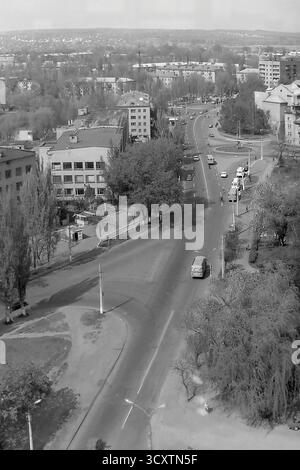Une photo d'archives panoramique à grand angle des années 1980 capture une scène urbaine animée à Sloviansk, en RSS d'Ukraine. La vue donne sur le carrefour où la rue Dobrovolskoho fusionne avec la rue Yunykh Kommunarov (maintenant rue Bankivska). La large avenue bordée d'arbres est remplie de voitures classiques de l'ère soviétique et d'un fourgon UAZ 'Bukhanka', mettant en valeur la circulation typique de la ville. Cet instantané du mouvement quotidien est un document historique précieux, préservant le rythme ordinaire de la vie dans un Donbass pacifique avant la guerre Banque D'Images