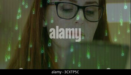 Regardant développeur féminin portant des lunettes analysant le code sur ordinateur portable dans l'étude, avec superposition binaire Banque D'Images