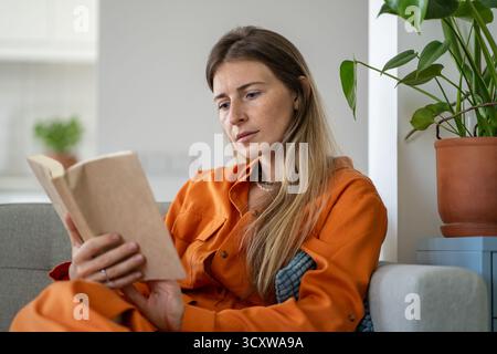 Femme sérieuse d'âge moyen lisant intentionnellement le livre de papier tout en étant assise sur un canapé confortable à la maison Banque D'Images
