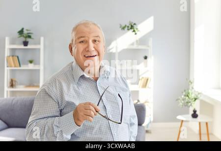 Portrait d'un homme âgé debout dans le salon et tenant des lunettes dans sa main. Banque D'Images