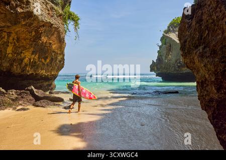 Surfeur va en mer à Uluwatu Beach (Blue point Beach), pittoresque plage de sable isolée entourée de falaises. Pecatu, Badung, Bali, Indonésie. Banque D'Images