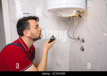 Plombier inspectant chauffe-eau avec lampe de poche dans la salle de bains Banque D'Images