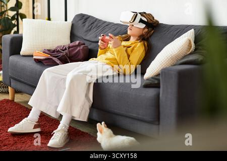 Une fille se détend sur le canapé, profondément engagée dans un monde de jeu virtuel tout en portant un casque. Banque D'Images
