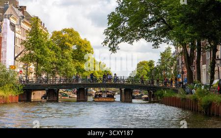 Amsterdam, pays-Bas - 5 août 2025 : les visiteurs traversent le pont pendant que les bateaux glissent sur le canal sous un ciel ensoleillé. Banque D'Images