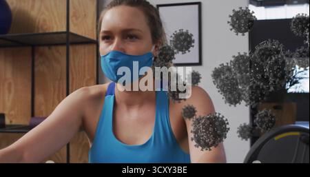 Femme portant débardeur bleu et masque facial pédalant vélo stationnaire dans la salle de gym à domicile, avec kettlebell Banque D'Images