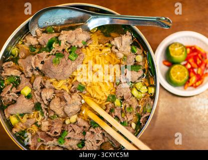 Cuisine traditionnelle du Vietnam, servie dans un bol en métal et une cuillère à soupe, avec des oignons verts frais, de l'ail et des nouilles de riz jaune dans un bouillon épicé, w Banque D'Images