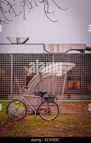 Bicyclette garée sous un abri transparent courbe. Un bâtiment en arrière-plan qui a une façade en forme de grille et des fenêtres. Banque D'Images