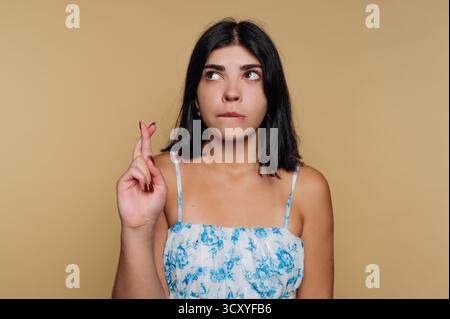 Une jeune femme aux cheveux foncés croise les doigts et regarde soigneusement, vêtue d'un haut floral clair. Le fond neutre ajoute un atmos calme Banque D'Images