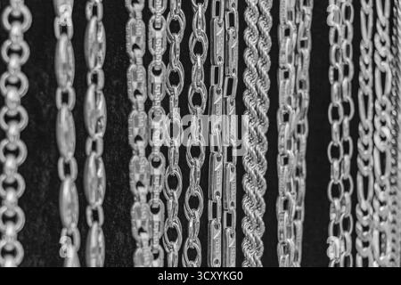 Un gros plan monochrome de diverses chaînes argentées suspendues en rangées sur un fond sombre. Chaque pièce affiche un savoir-faire fin et un motif de lien complexe Banque D'Images