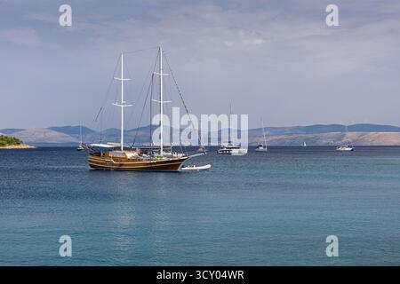 Port côtier tranquille avec un bateau à voile en bois classique ancré près d'un petit bateau gonflable, avec des collines lointaines et d'autres yachts parsemés t Banque D'Images