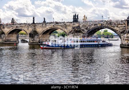 Bateau d'excursion passant sous le pont Charles sur la rivière Vltava à Prague, en République tchèque, avec des foules marchant sur le pont historique un jour d'été. Banque D'Images