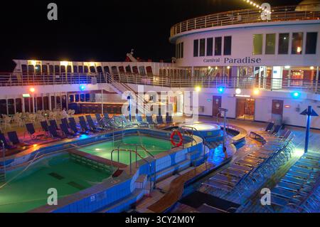 Vue nocturne sur le pont de la piscine du bateau de croisière Carnival Paradise illuminé par des lumières colorées, avec chaises longues et atmosphère calme en mer Banque D'Images