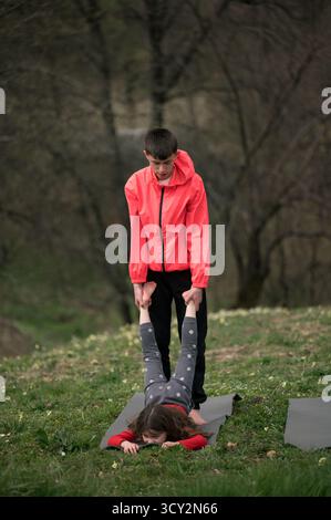 Deux jeunes individus s'engagent dans une pose de yoga partenaire dans un paysage verdoyant luxuriant, démontrant le travail d'équipe et l'équilibre sous la lumière chaude du soleil pendant un pois Banque D'Images